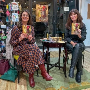Two women sitting in a bookstore, holding books, with bookshelves and a small table between them.