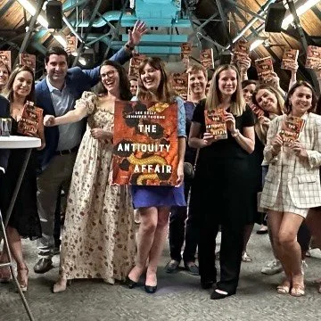 Group of people at a book launch celebration holding copies of a book titled 'The Anticority Affair' in a studio setting with lights.