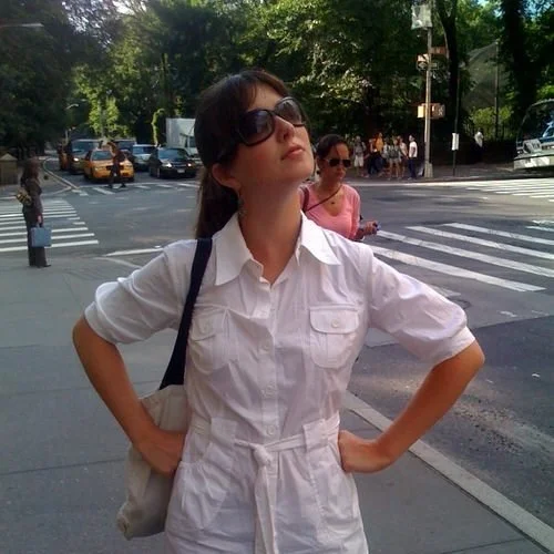 A young woman wearing sunglasses and a white jumpsuit, standing on a city street with her hands on her hips, during the daytime.