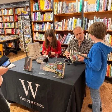 A woman and a man signing books at a table in a bookstore, with a young boy browsing books nearby. Bookshelves filled with books are in the background.