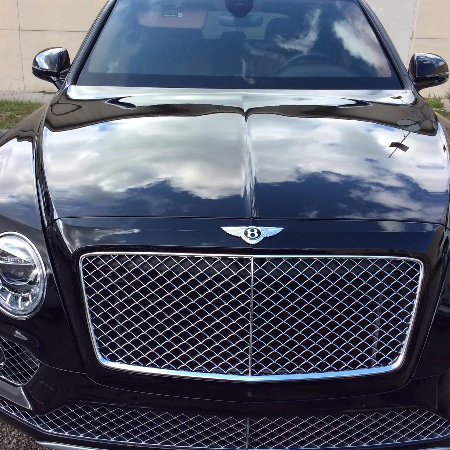 Front view of a black Bentley luxury car with a shiny chrome grille and Bentley emblem on the hood, reflecting the sky and clouds.