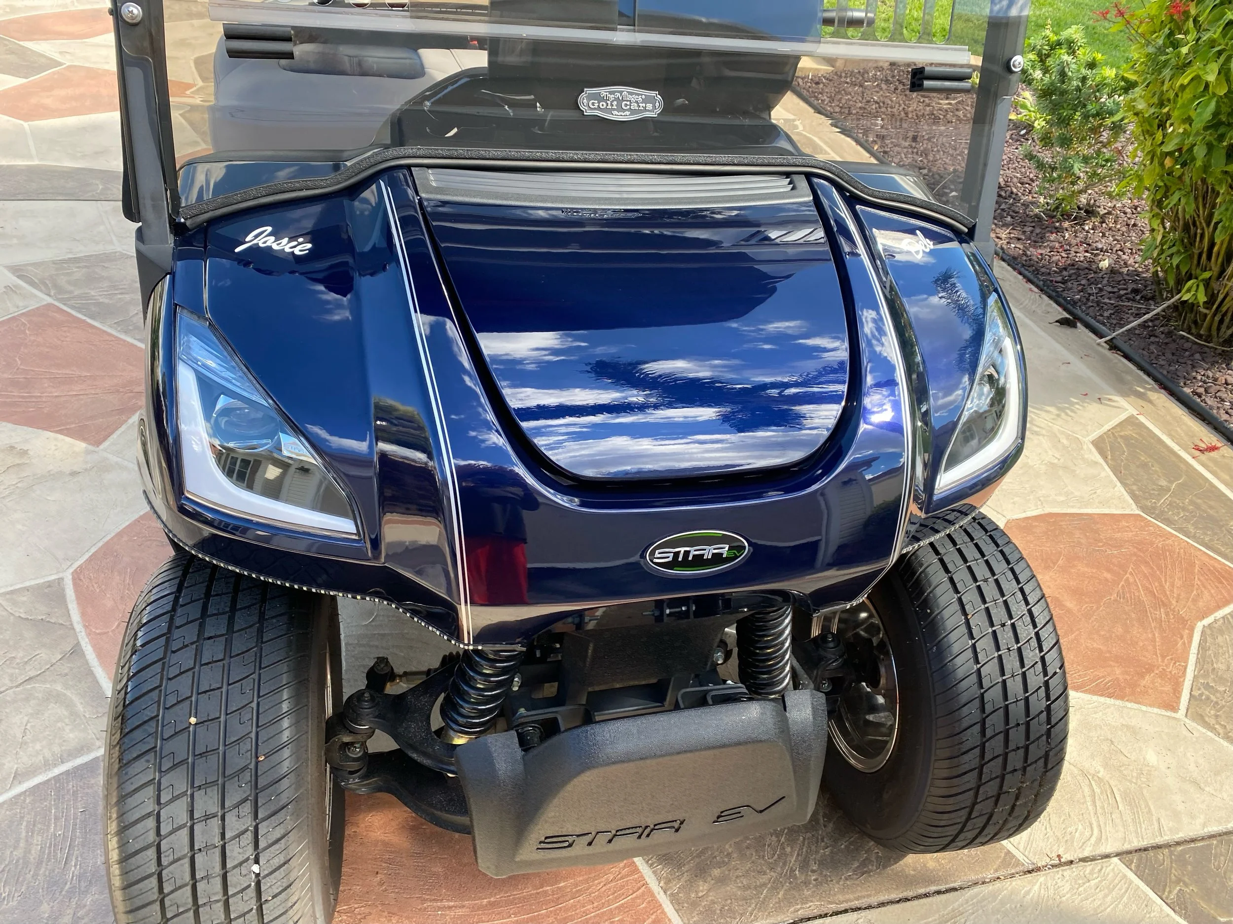 Close-up of a blue golf cart with the name 'Josie' on the front. The cart has a black and silver logo that reads 'The Village Golf Cars' on the windshield. The front of the golf cart features large tires, suspension, and a black chassis.