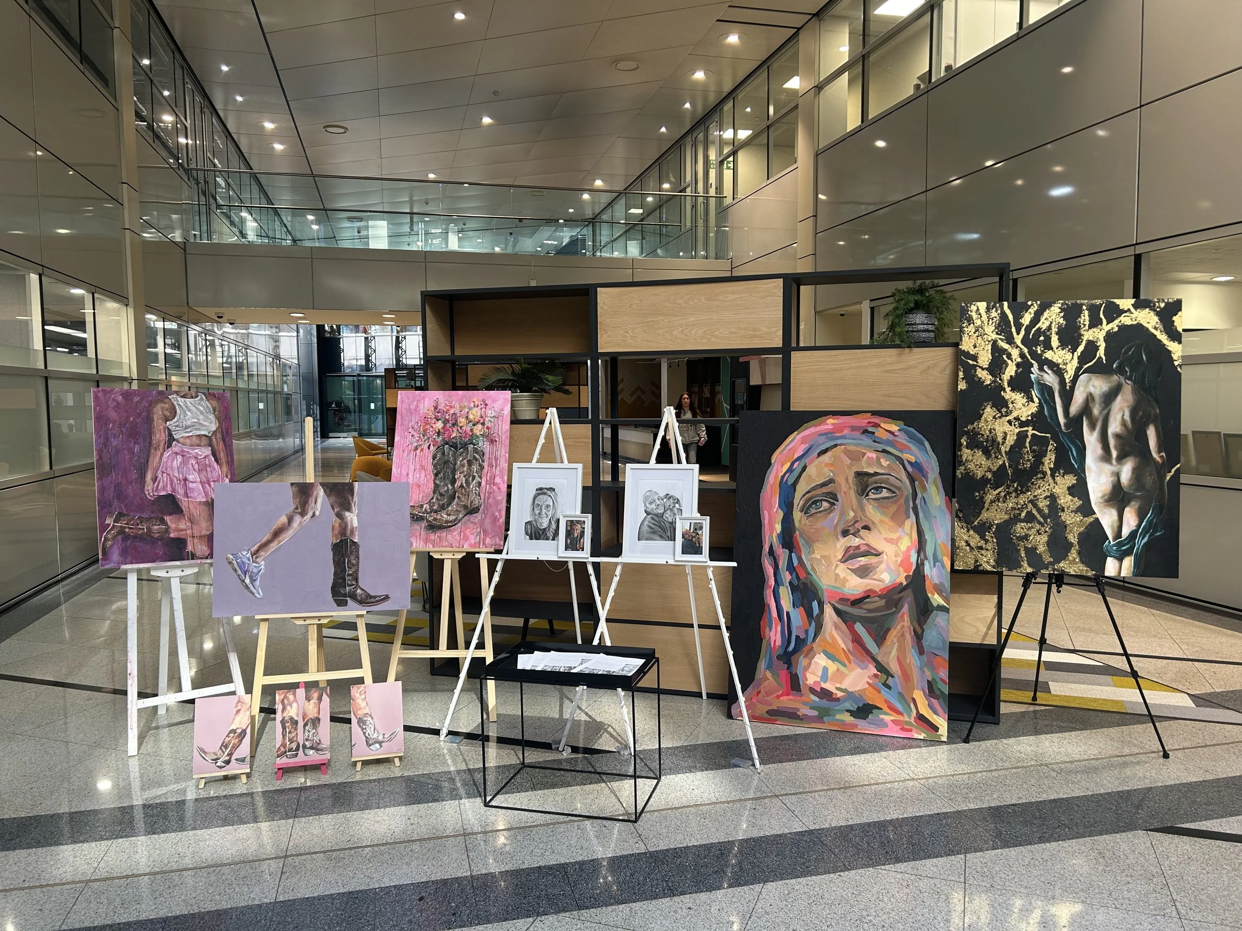 Art exhibit displaying paintings, including a colorful portrait of a woman with braided hair, a girl in a pink dress, and other artistic portraits, set up in a modern indoor space with glass walls and multiple lights.