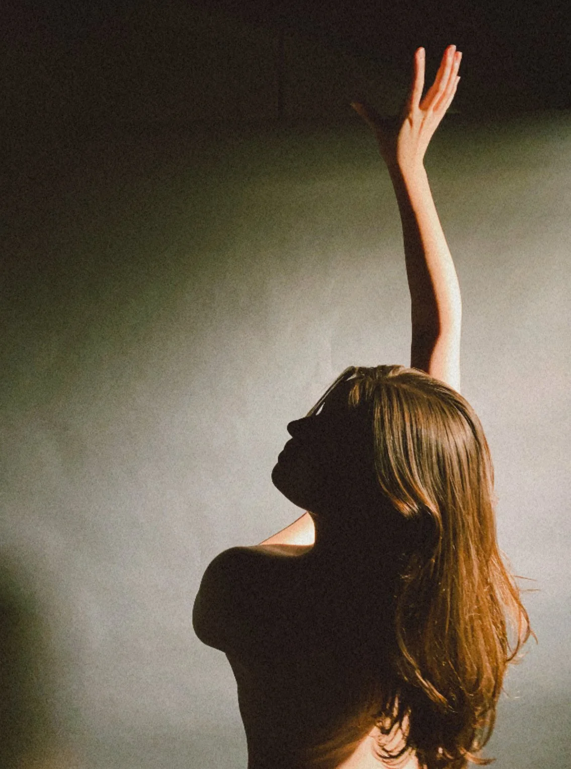A silhouette of a woman with long hair posing against a neutral background, with one arm raised above her head.