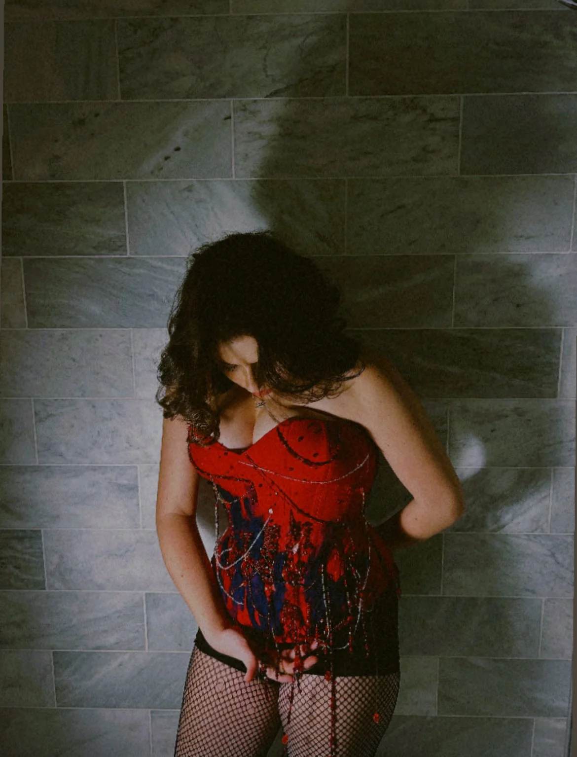A woman with curly brown hair wearing a red, black, and blue dress with fishnet stockings, standing against a gray stone wall.