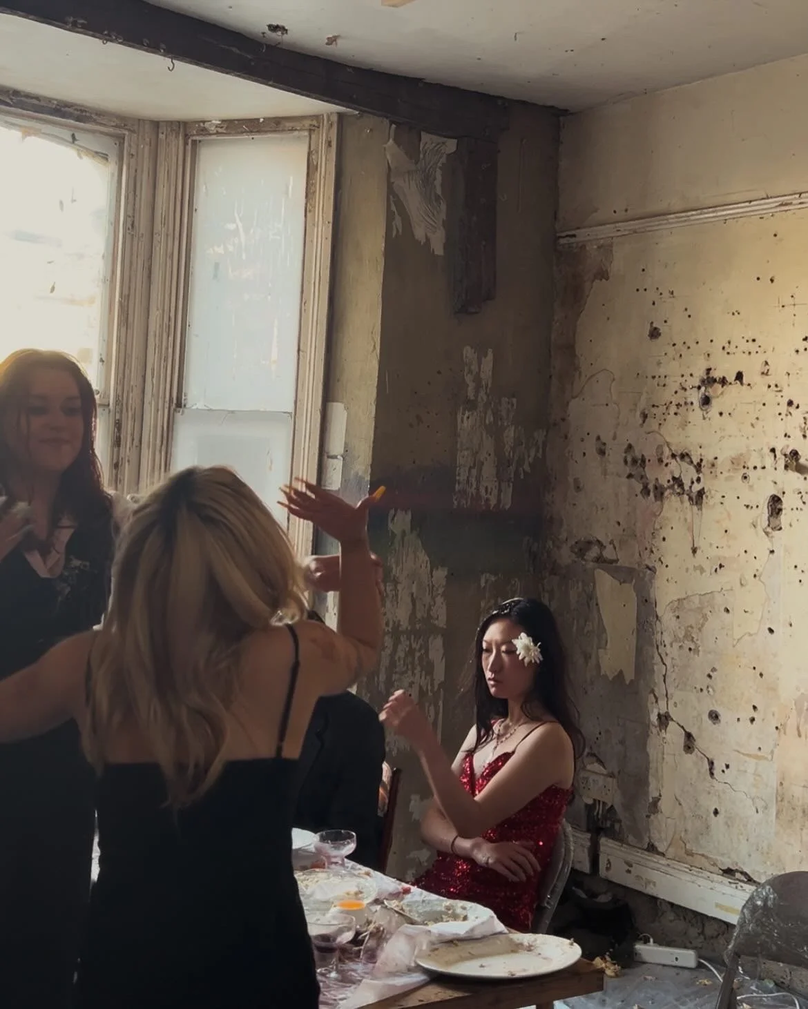 Young woman in a red sequined dress sitting with her eyes closed while others converse at a damaged, peeling wall room with a table of scattered food.