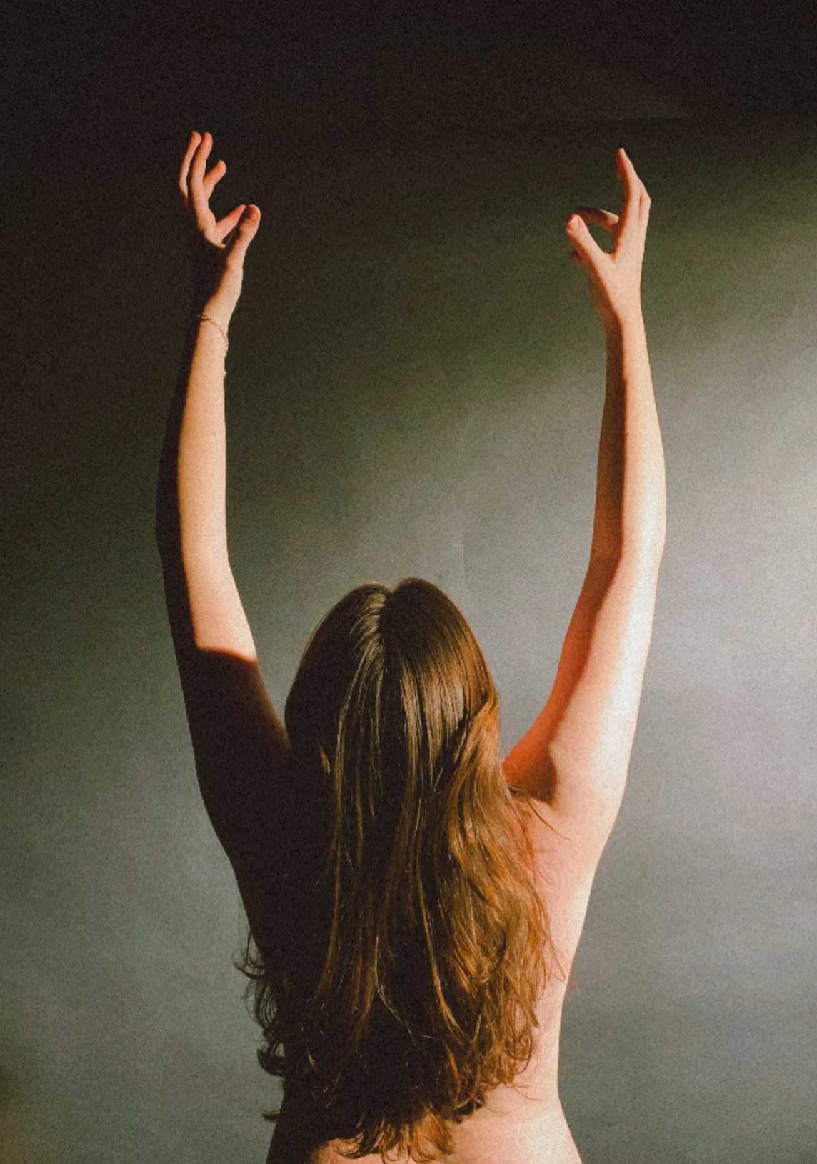 A woman with long brown hair raising her arms above her head against a dark background.
