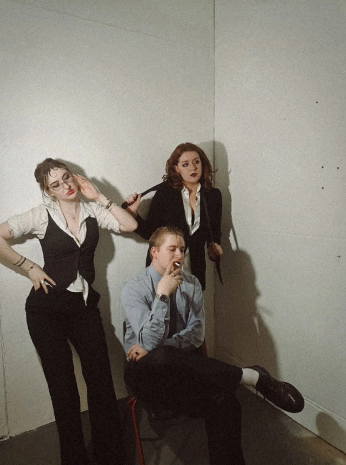 Three young adults posing against a plain white wall: a woman with glasses and dark lipstick on the left, a woman with curly hair and dark lipstick in the center, and a man sitting on a chair smoking a cigarette in the foreground.