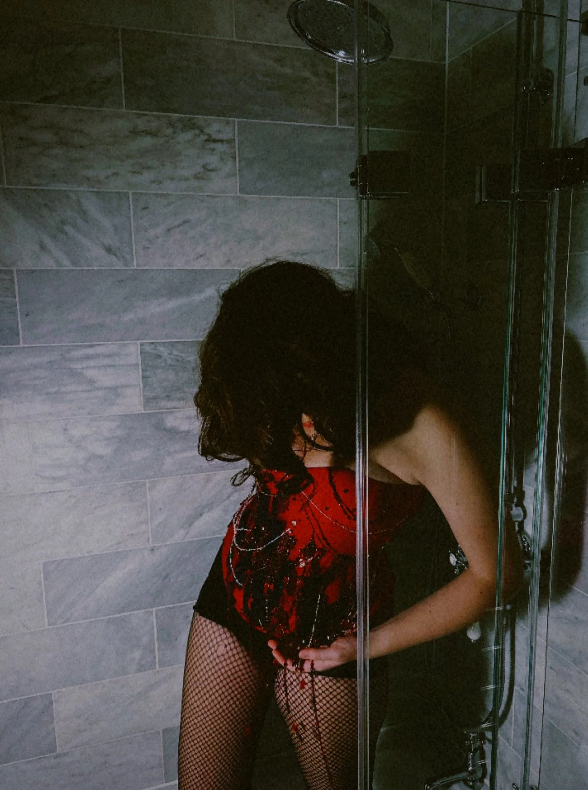 Person with black hair in a red and black outfit and fishnet stockings taking a mirror selfie in a tiled bathroom shower