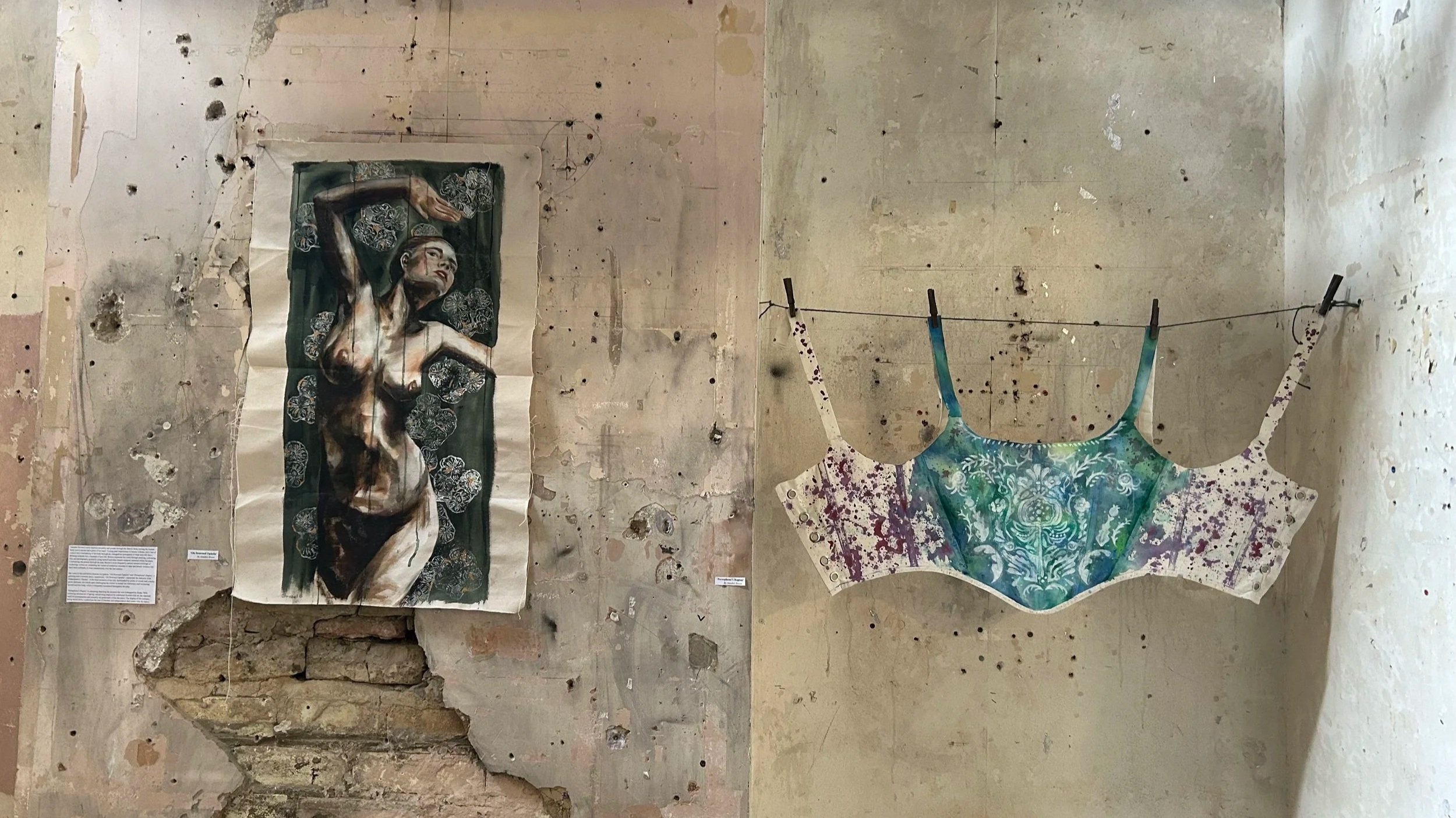 A wall with holes and peeling paint, featuring three pieces of art: a realistic painting of a woman with dark hair and an arm raised, and two dresses hanging from a clothesline.