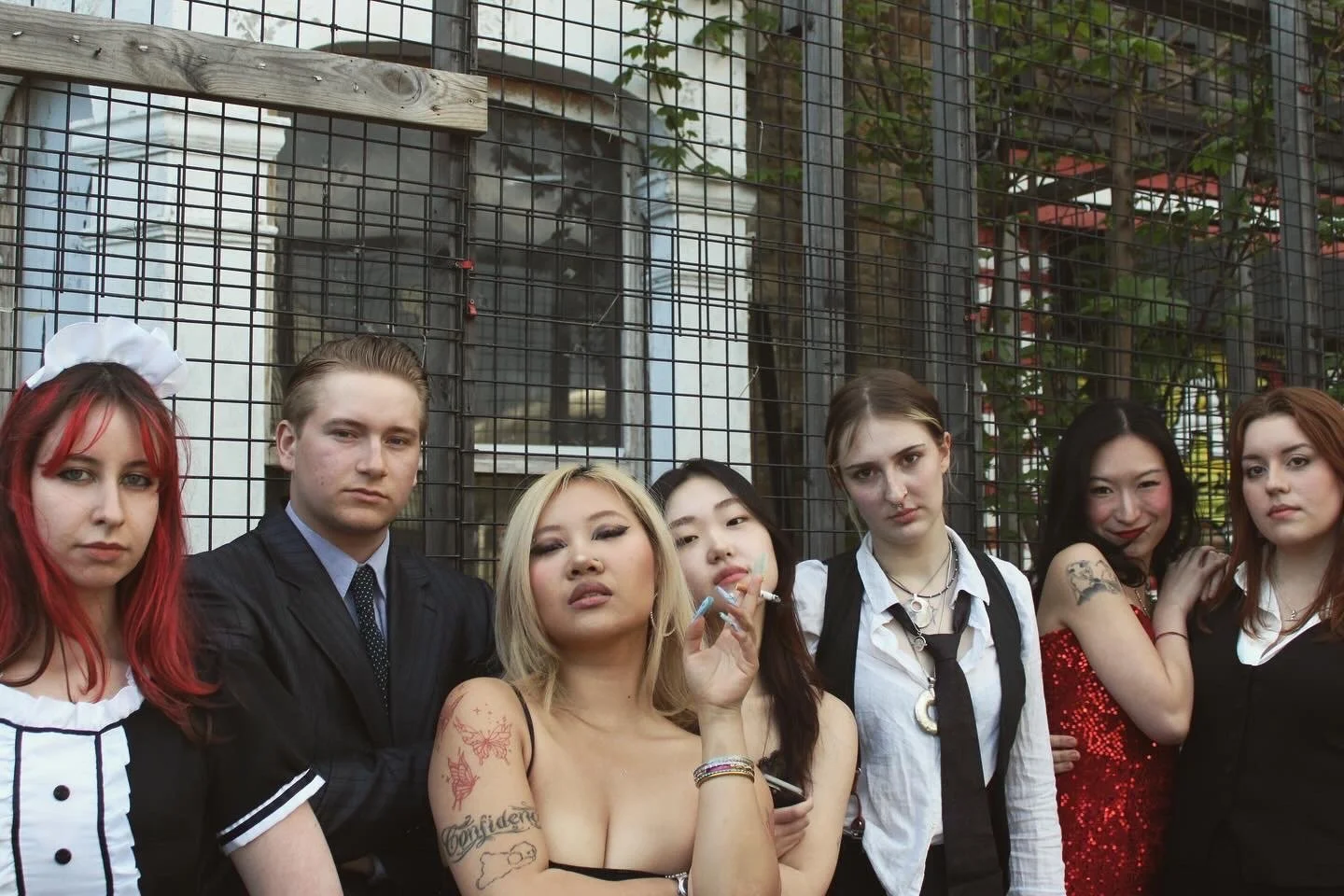 Group of six diverse young women and one young man standing in front of a wire fence, dressed in alternative and vintage styles, posing confidently.