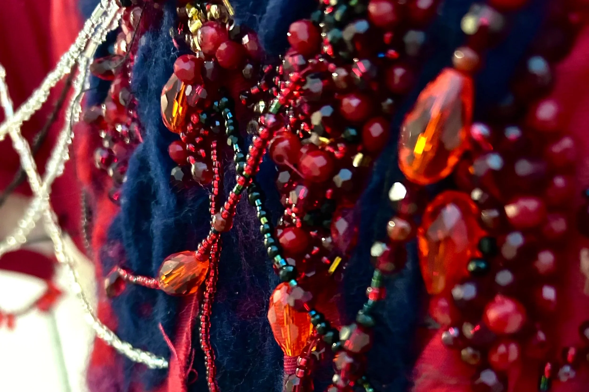 Close-up photo of various red, orange, and multicolored shiny beads and jewelry, possibly necklaces or bracelets, on a dark blue fabric background.
