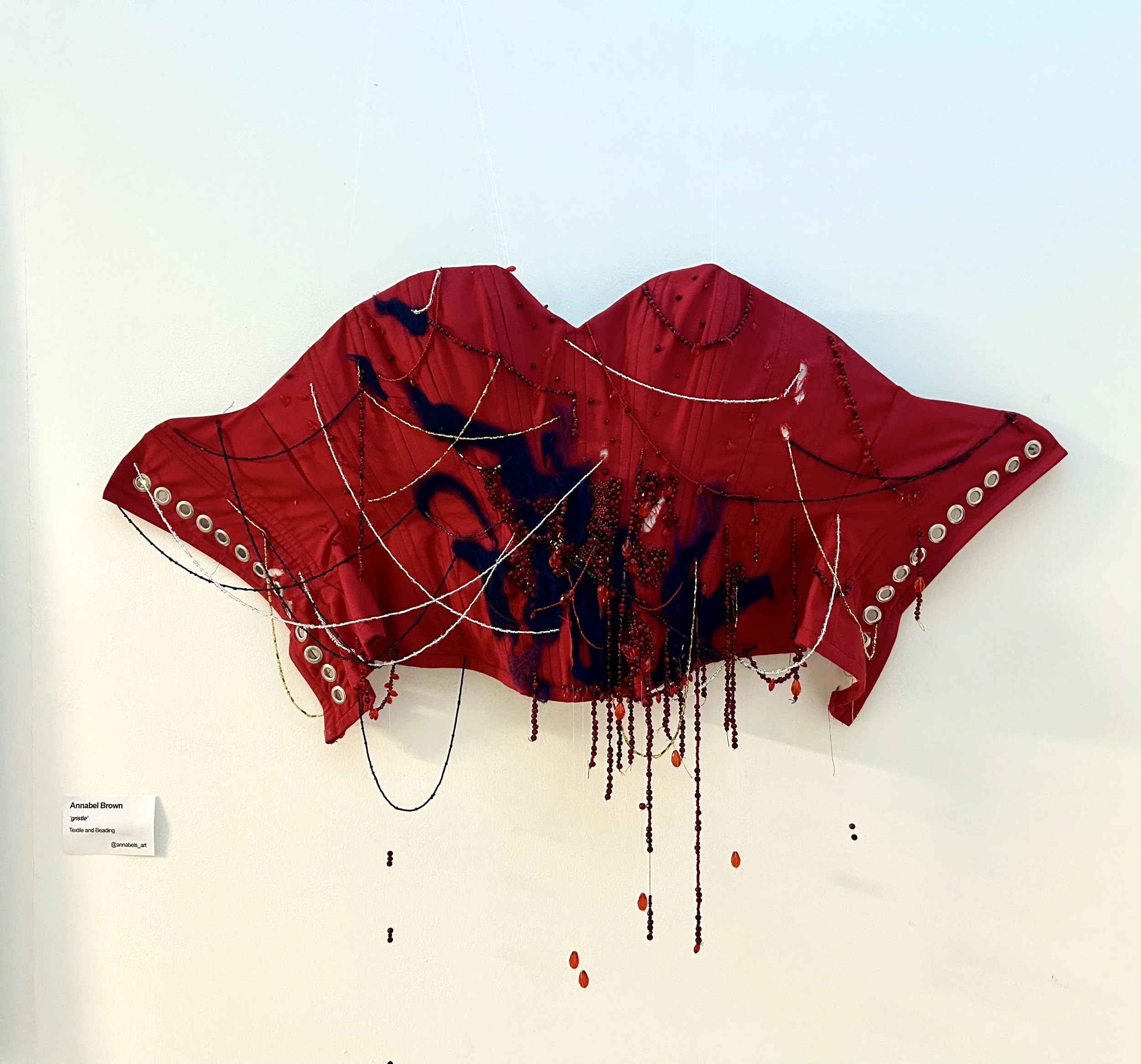 Red textile art piece with black and red beading and hanging beads, displayed on a white wall.