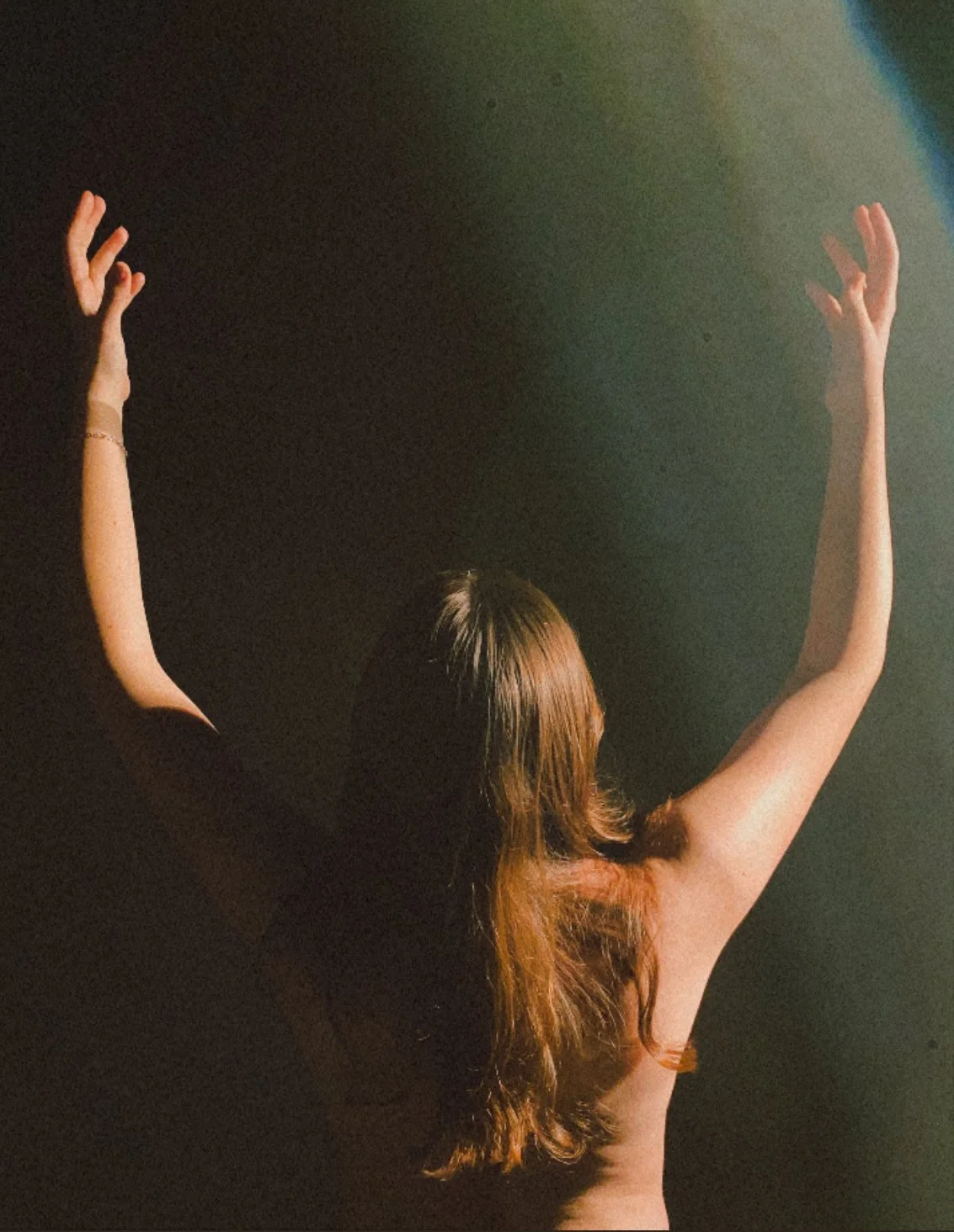 A woman with long, brown hair standing against a dark background with her back to the camera, raising her arms in the air with her fingers spread apart.