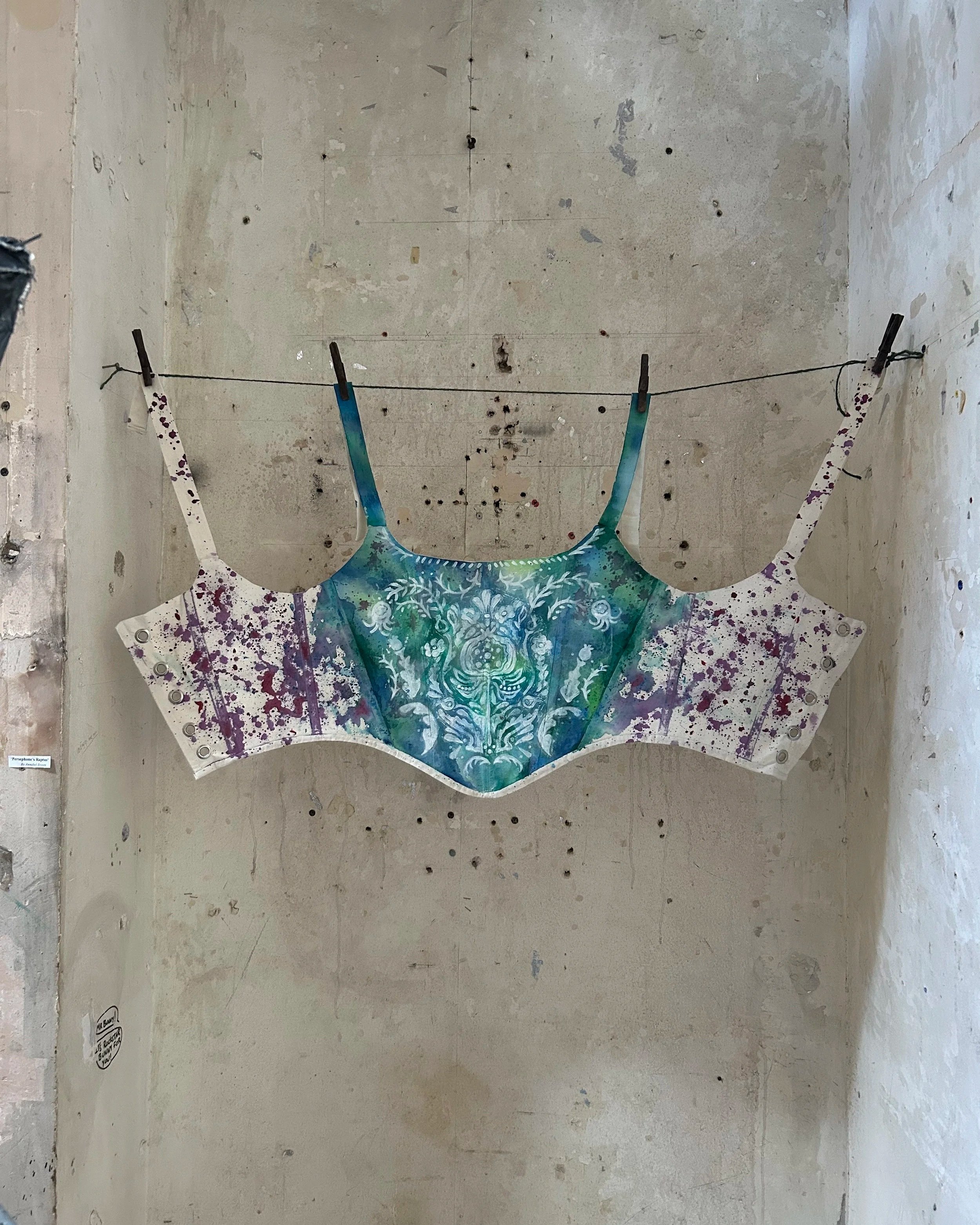 Colorful abstract art piece hanging on a gray, worn concrete wall with holes and marks. It is attached with clothespins on a string.