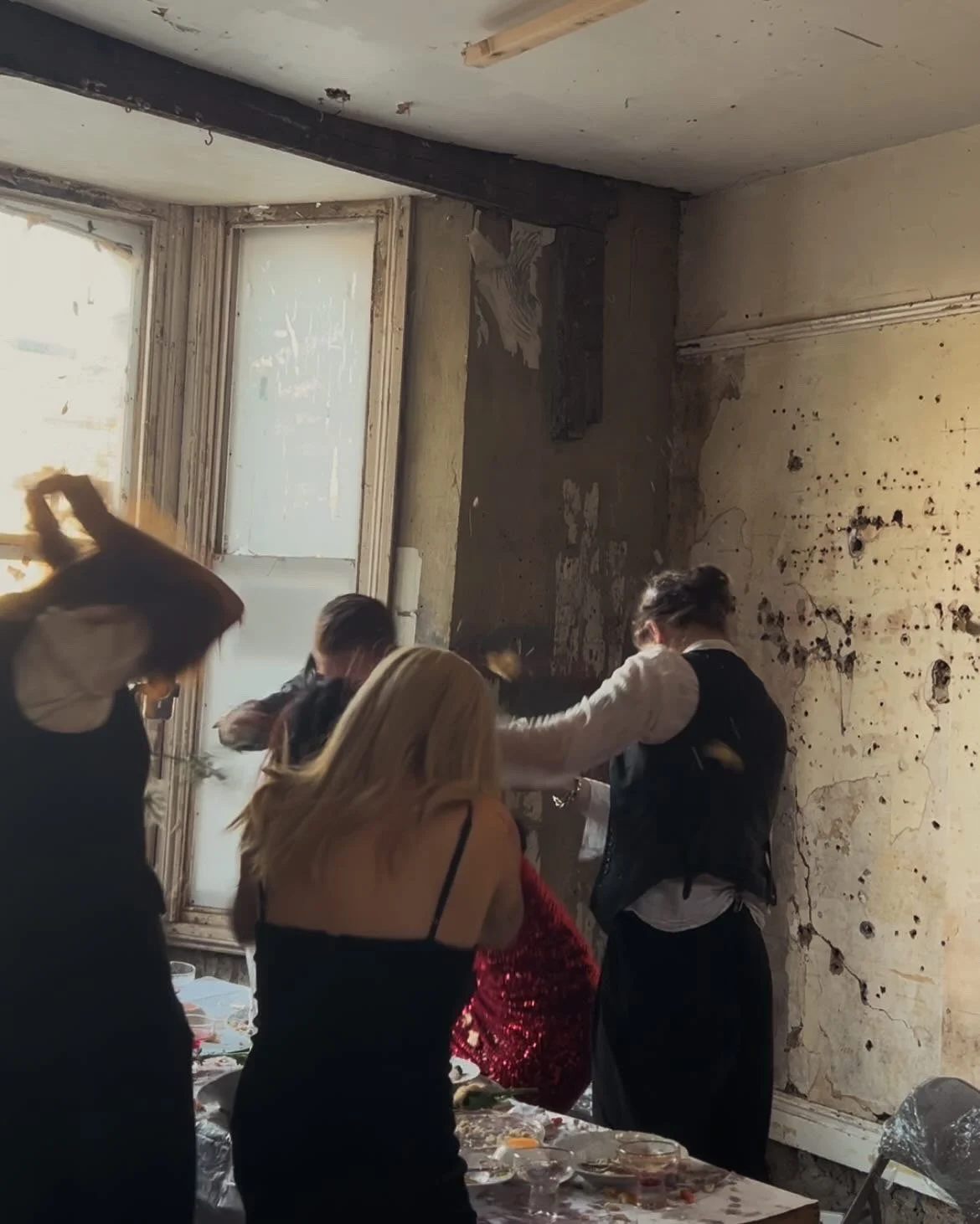 People in formal attire gather around a table in a room with peeling paint and damaged walls, possibly for a celebration or event.