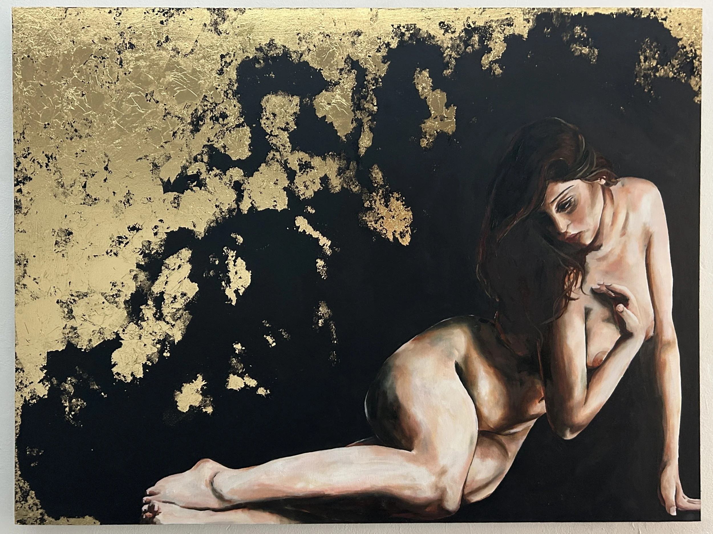 A painting of a nude woman with long dark hair, sitting with her knees drawn up and her arms crossed over her chest, against a black background with gold leaf accents.