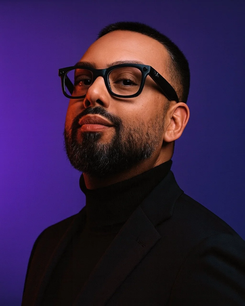 A portrait of EMKWAN with glasses, a beard, and short hair, wearing a black turtleneck and blazer, against a purple background.