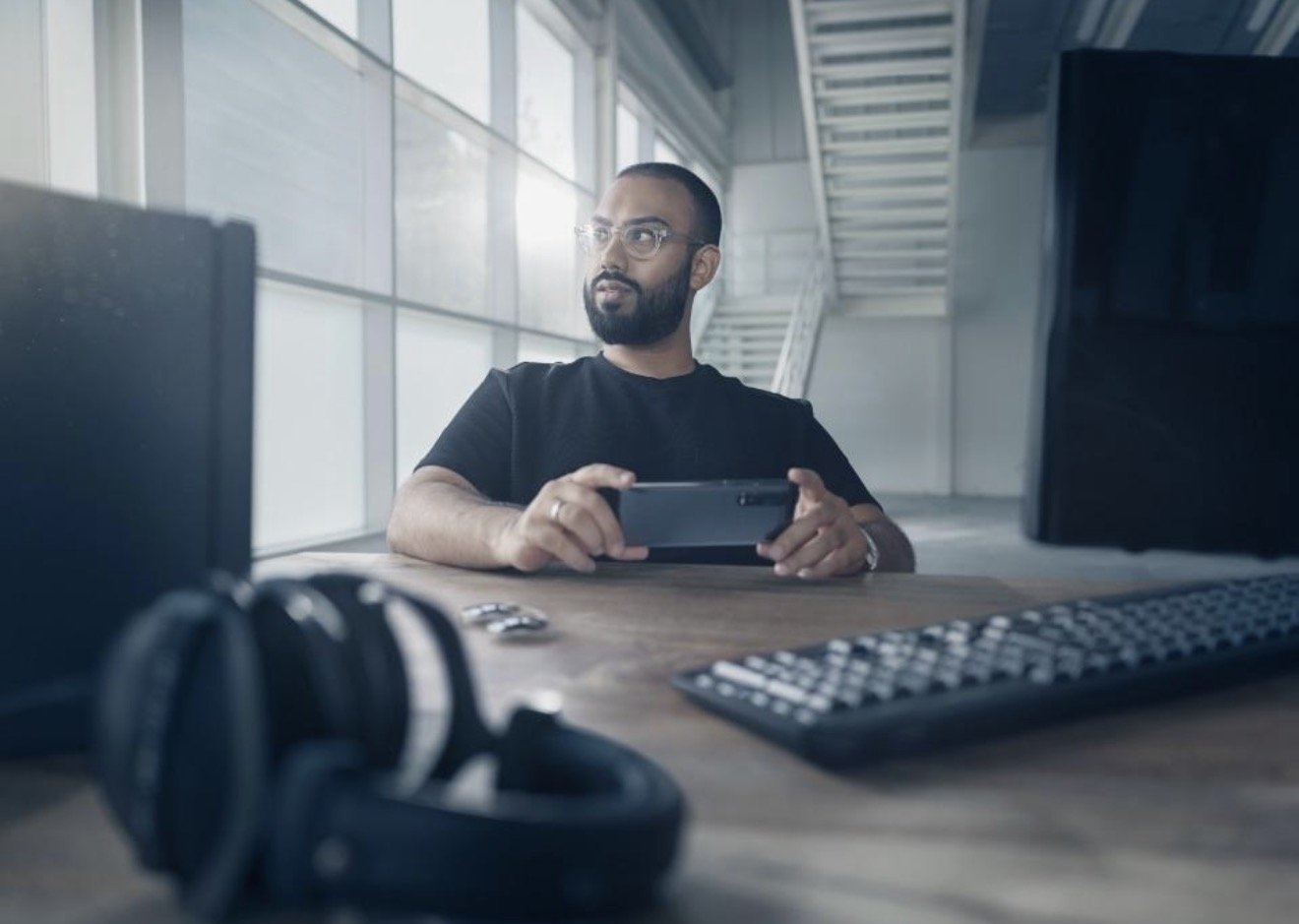 Emkwan sitting at a desk in a modern office, holding a smartphone, with computer monitors, keyboard, and headphones on the desk. Large windows in the background.