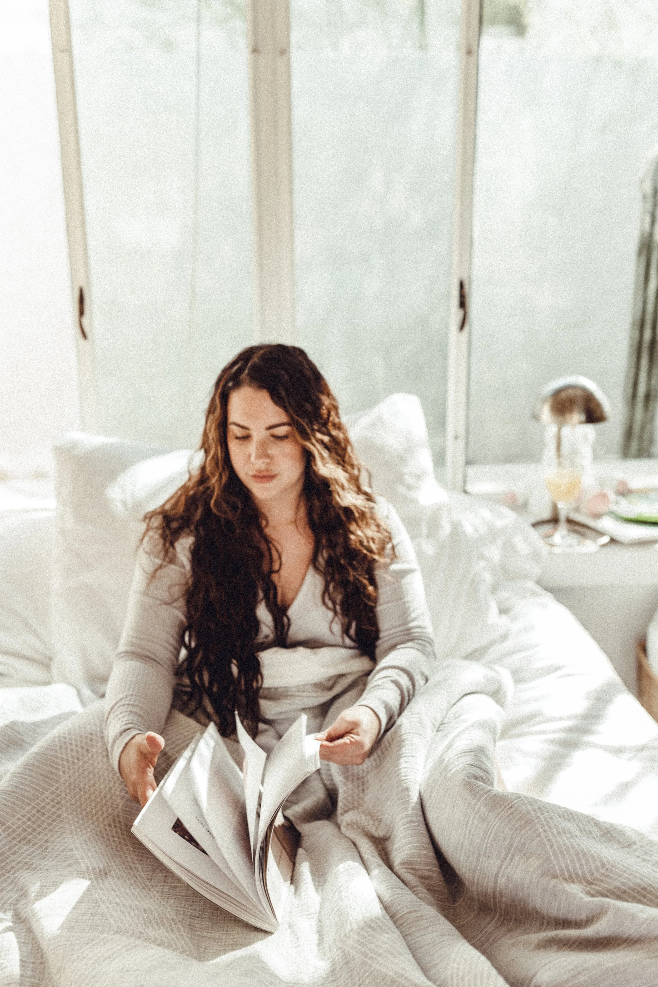 A woman with long, wavy hair sitting on a bed, reading a magazine in a bright bedroom with white bedding and a nightstand with a lamp.