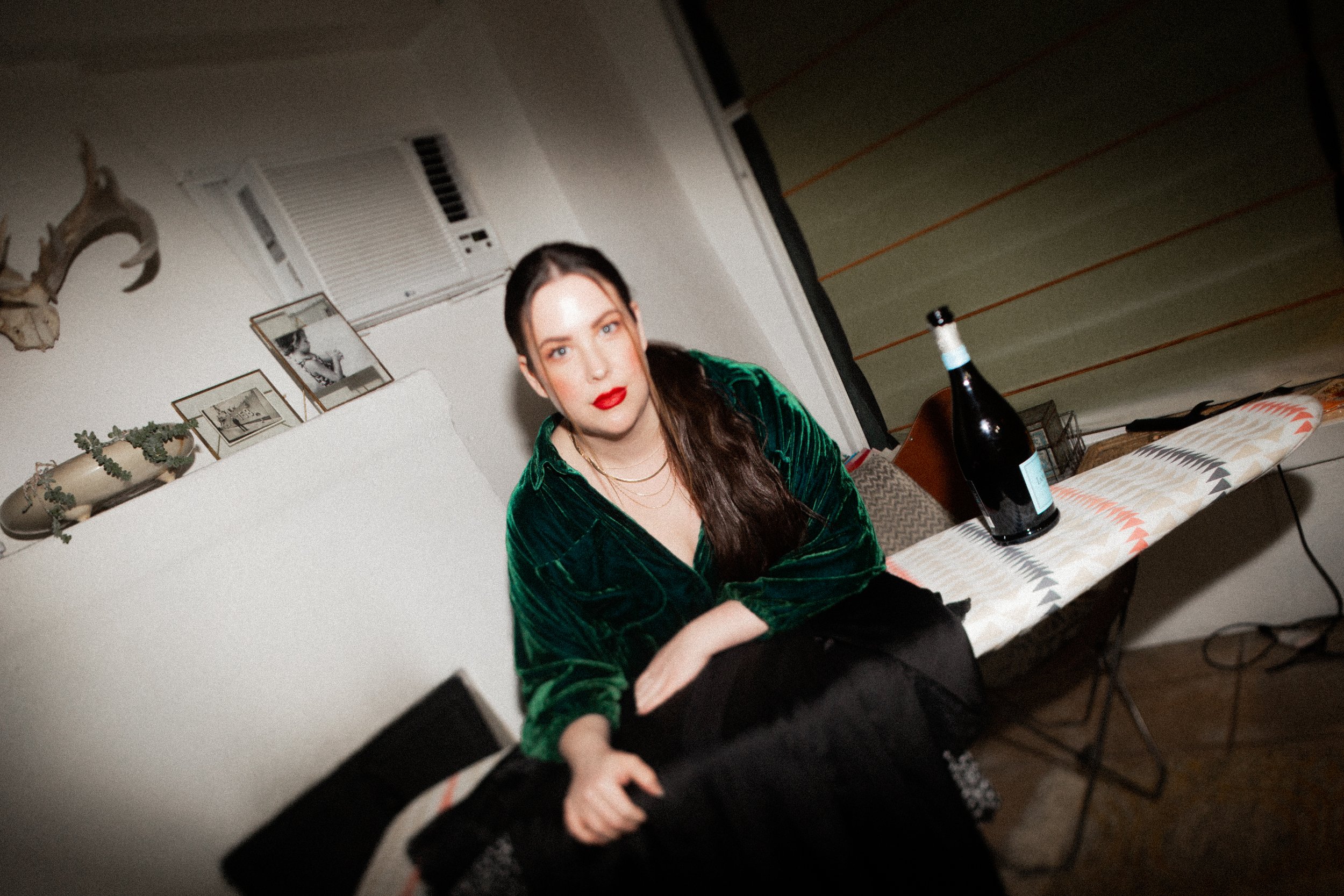 A woman with long 24 inch extensions dark hair, red lipstick, and blue eyes wearing a green velvet top sitting at a table with a champagne bottle. This is a photoshoot styled and shot for Aloe Studio.