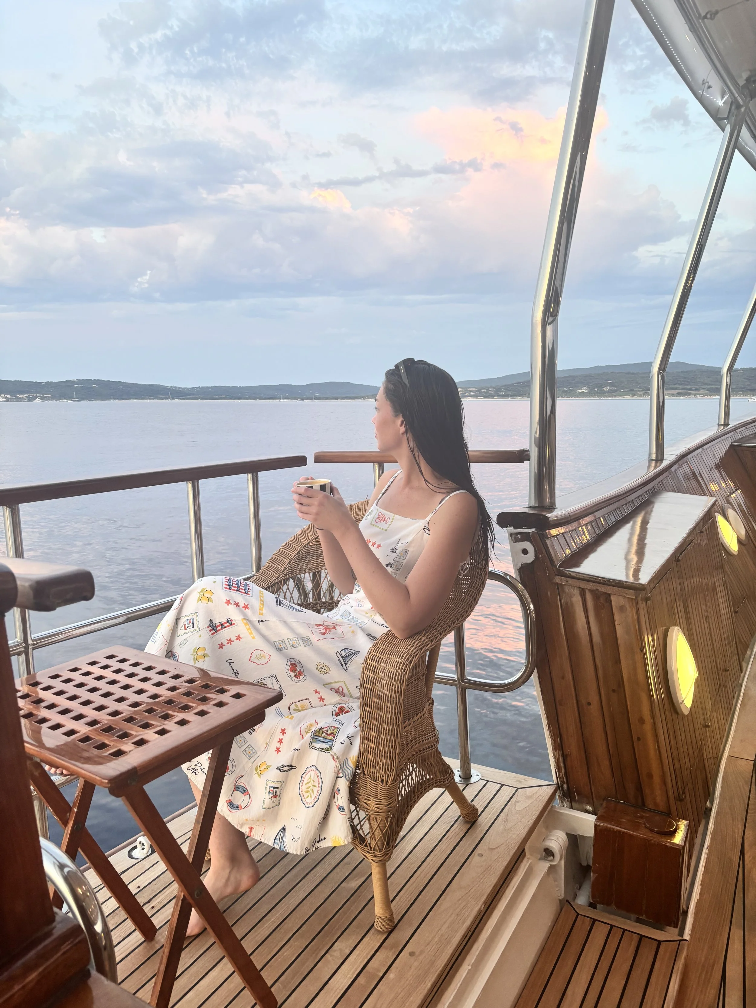 A woman sitting on a wicker chair on a boat deck, holding a cup, with water and distant hills visible in the background during sunset.