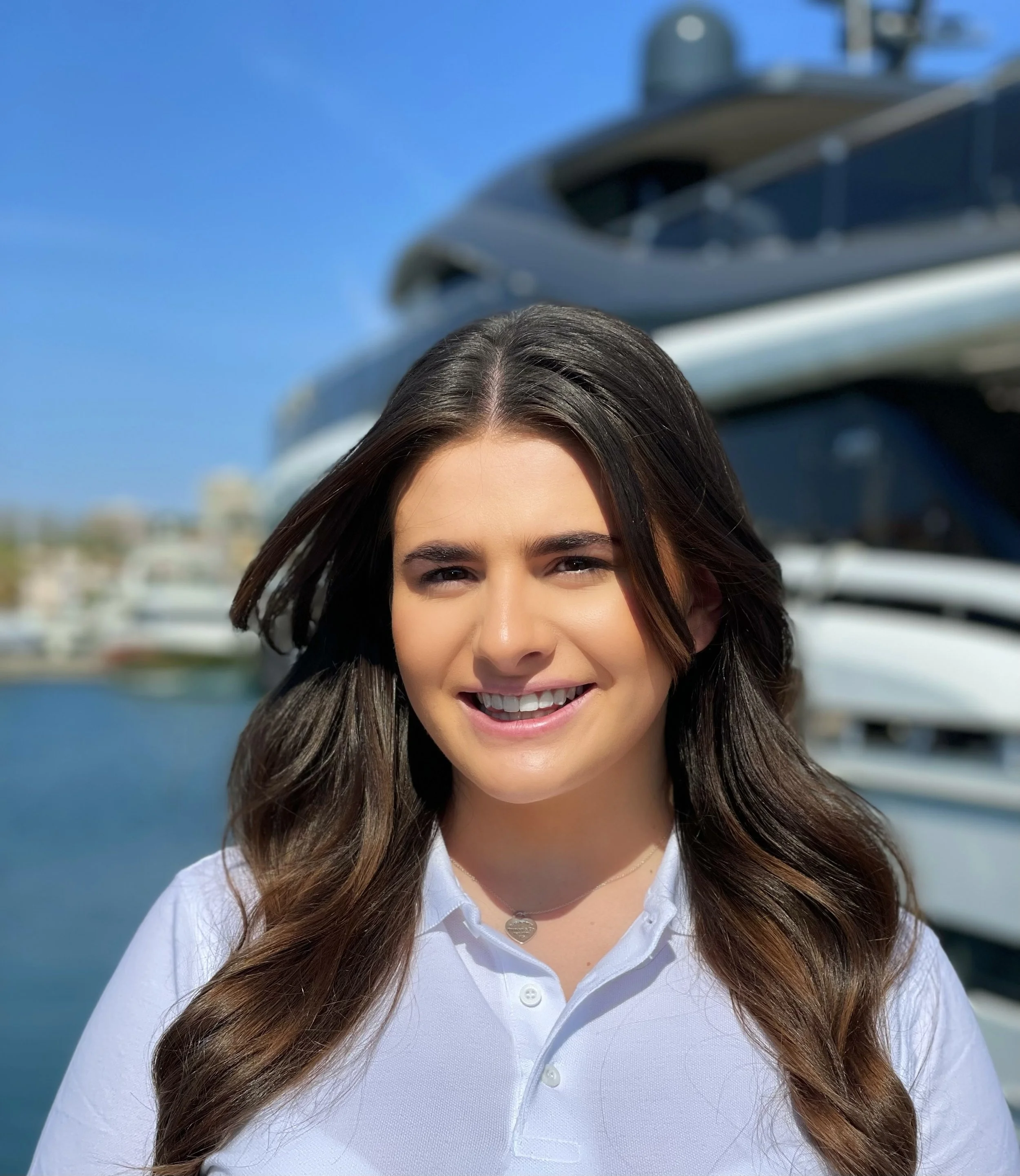 Close-up of a smiling woman with long brown hair, wearing a white polo shirt and a heart-shaped necklace, outdoors with a yacht and blue sky in the background.