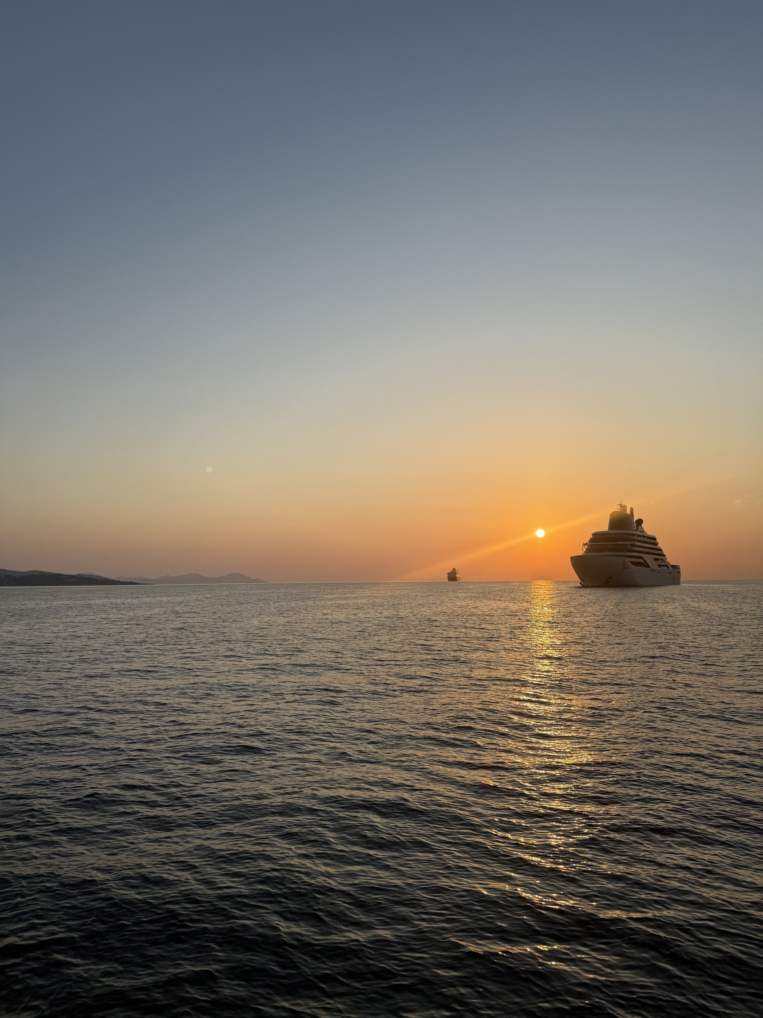 A large super yacht sailing on the calm ocean during sunset with the sun near the horizon, casting a golden glow on the water, and a smaller ship visible in the distance.
