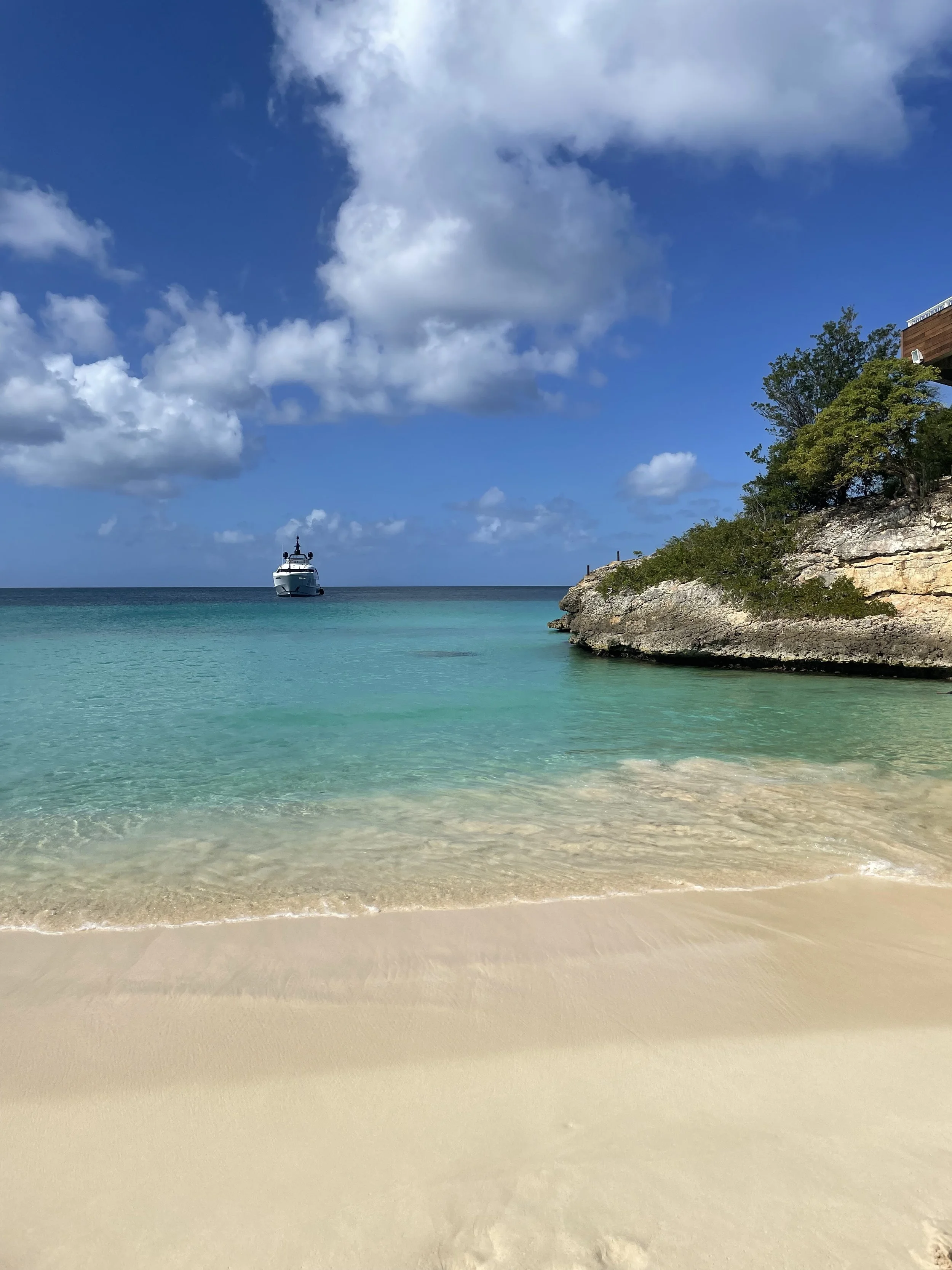 A beach with clear turquoise water, white sand, a rocky outcrop with trees on the right, and a yacht anchored in the distance under partly cloudy skies.