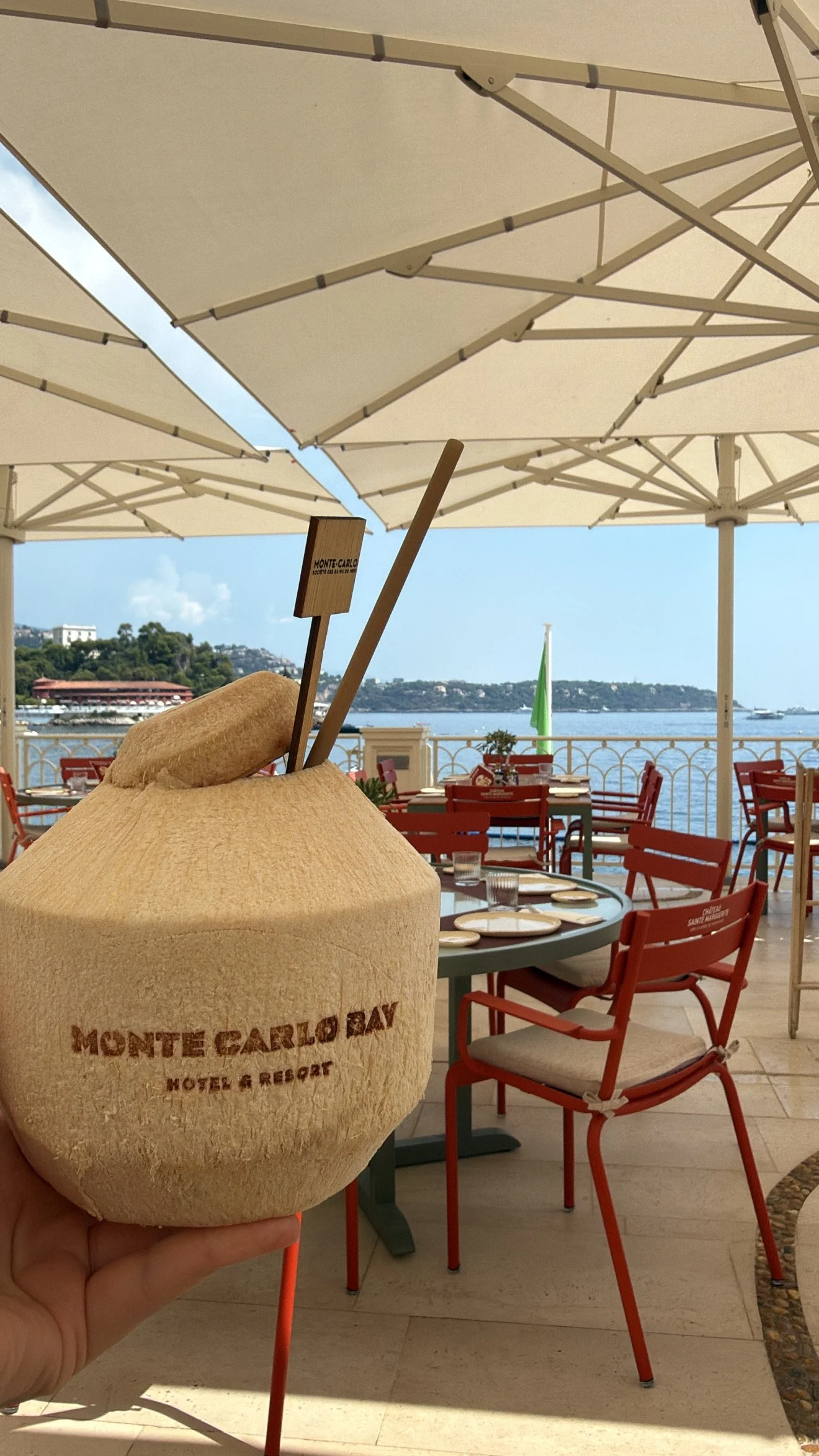 A beach club or restaurant at Monte Carlo Bay Hotel & Resort, with outdoor seating, red chairs, tables set with glasses and plates, large white umbrellas, and water in the background.