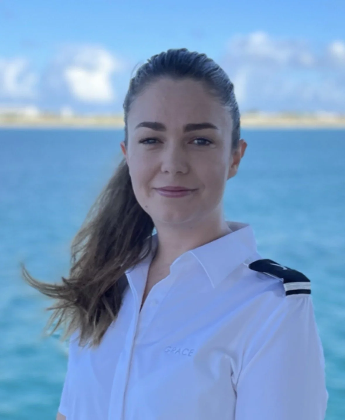 Young woman in white uniform with shoulder patches smiling by water with a distant shoreline and blue sky in the background.
