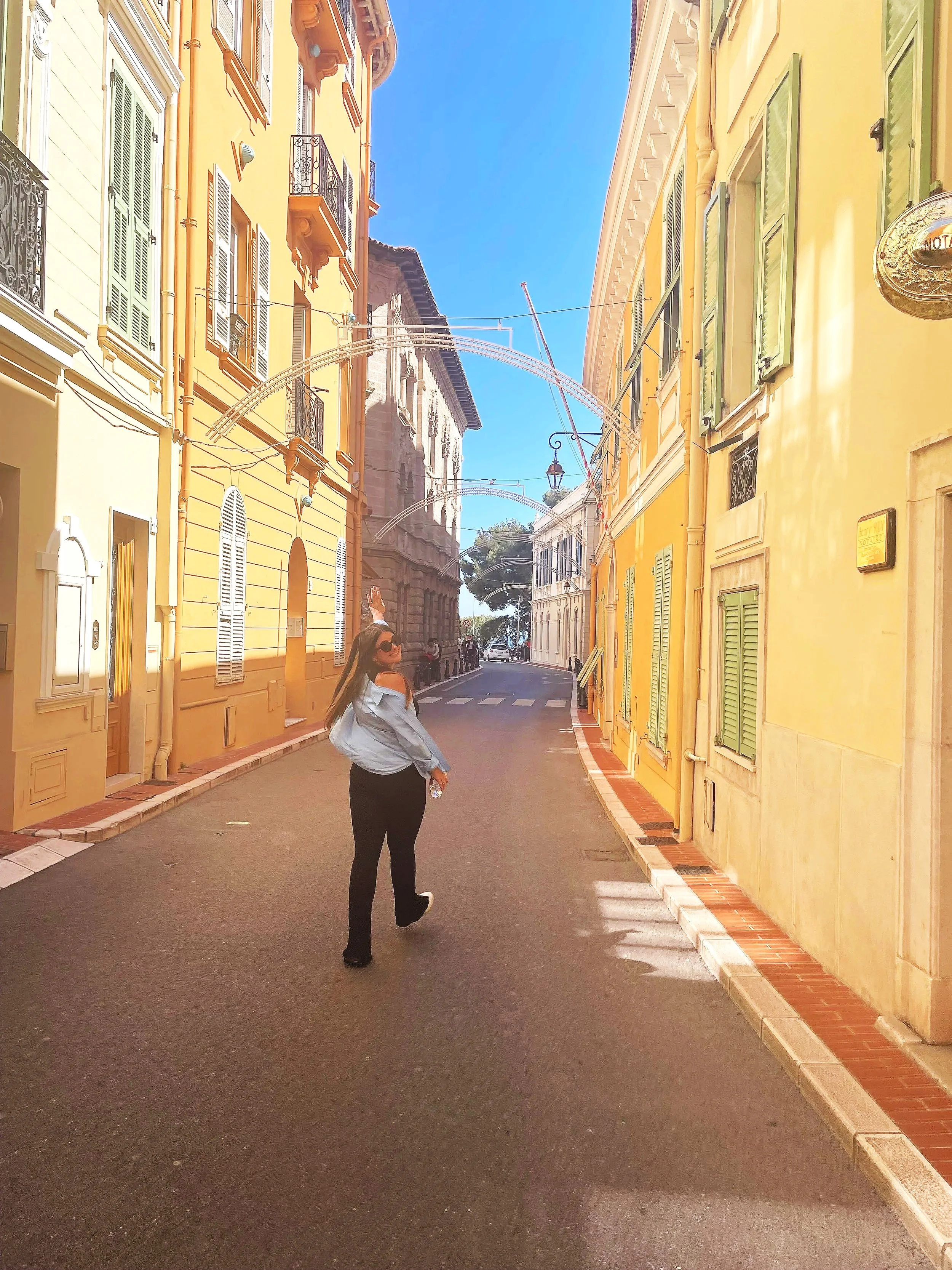 A woman in sunglasses, a white jacket, and dark pants standing in a narrow street lined with colorful buildings with green shutters.