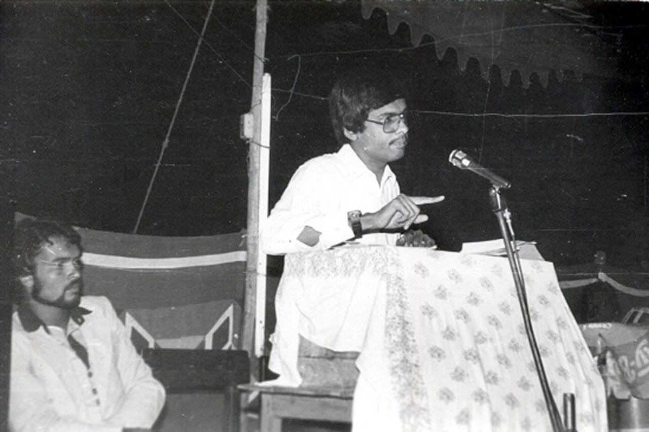 A man with glasses speaking at a podium covered with a floral cloth, gesturing with his hands, while another man sits nearby.