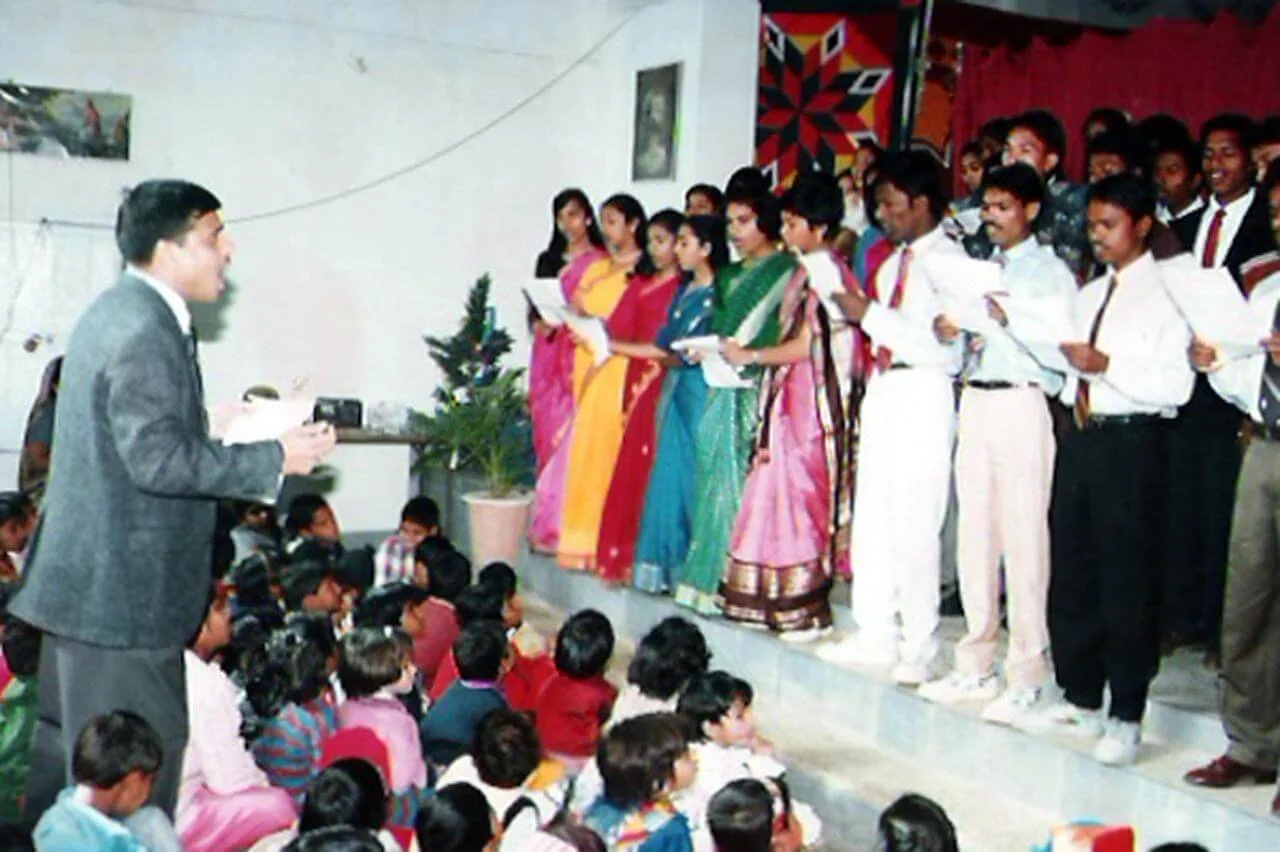 A school concert with students singing on stage, dressed in formal attire, with an audience of children sitting on the floor.