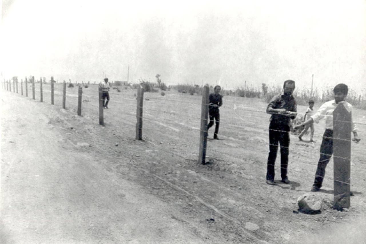 Children playing near a barbed wire fence on a dirt field in a rural area.