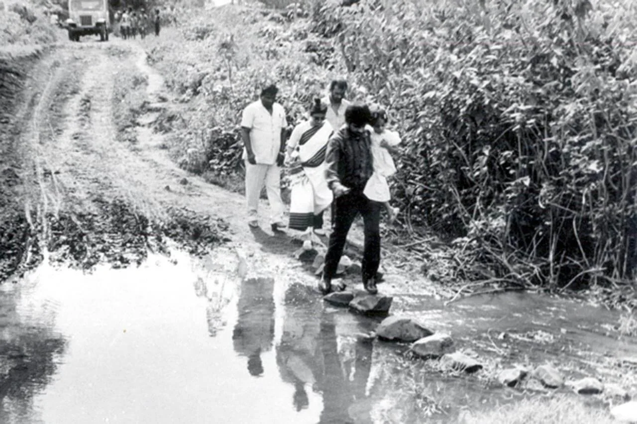 Four people walking on a stone pathway beside a stream through a natural setting, with a vehicle in the background.