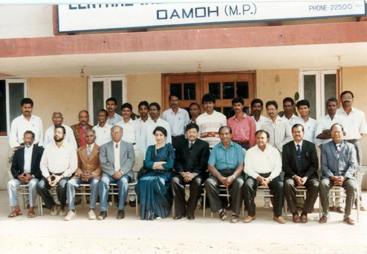 Group of around 22 people, mostly men, sitting and standing in front of a building with a sign in a non-English script, possibly a formal gathering or meeting.