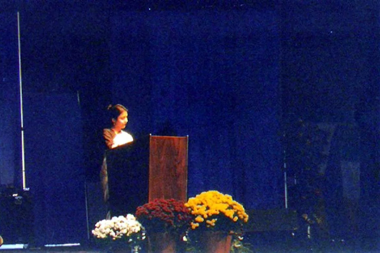 A woman standing behind a wooden podium on a stage decorated with yellow, orange, red, and white flowers, with a dark blue curtain in the background.
