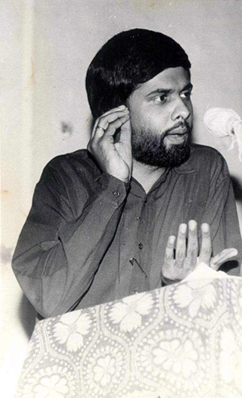 Black and white photo of a man with a beard and dark hair, wearing a button-up shirt, gesturing with his hands, seated at a table with a patterned cloth, and speaking into a microphone.