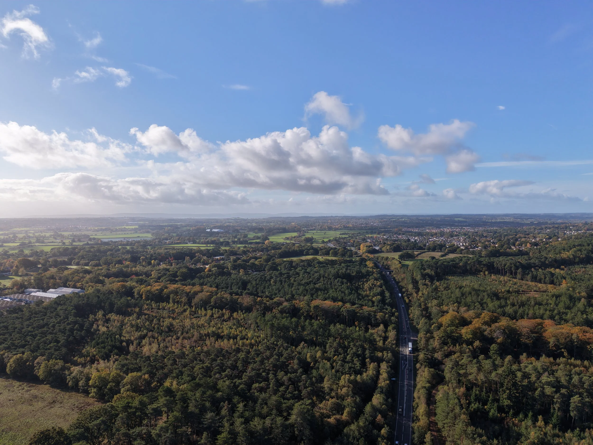 Aerial view of a forested landscape, winding road, green fields, and suburban neighborhoods beneath a blue sky with scattered clouds.