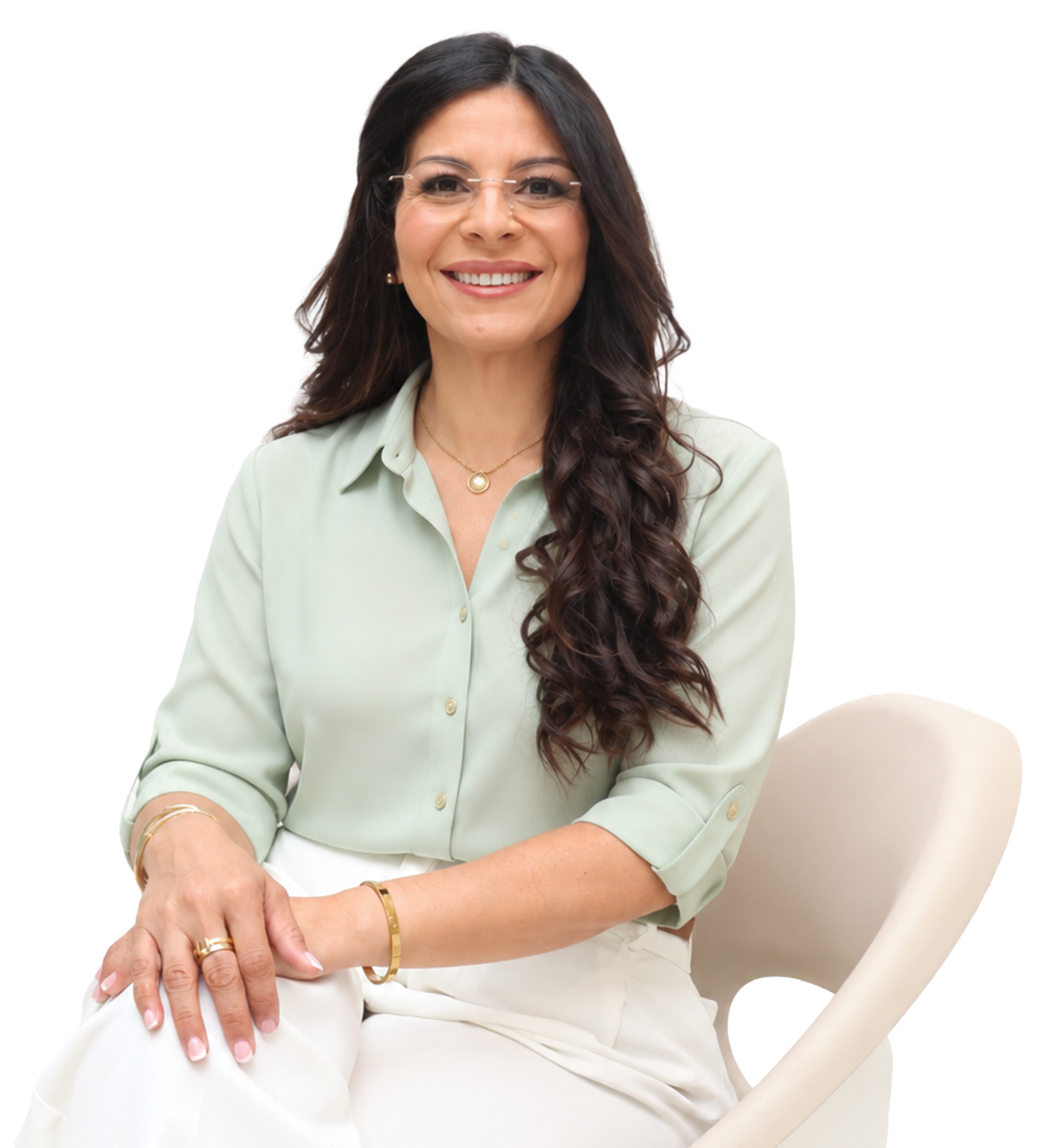Mujer joven con cabello castaño oscuro y rizado, usando gafas, camisa de color crema y accesorios dorados, sonriendo, sentada en una silla blanca.