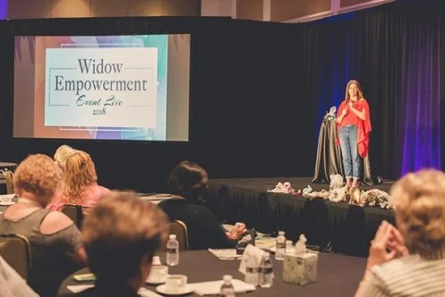 A woman speaking on stage at the Widow Empowerment Event Live 2018, with a large screen displaying the event name behind her, in a dimly lit conference room with attendees seated at tables.