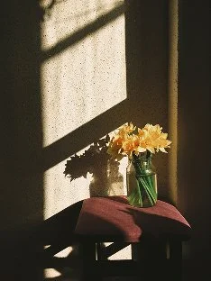 A glass jar with yellow flowers on a small table with a purple cloth, sunlight casting shadows inside a room.