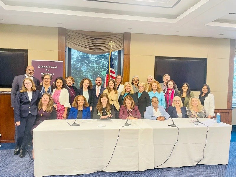 Group of women and a few men in a conference room with an American flag, a banner reading 'Global Fund for Women,' and a large window with trees outside. Some people are seated at a table with microphones, others are standing behind.