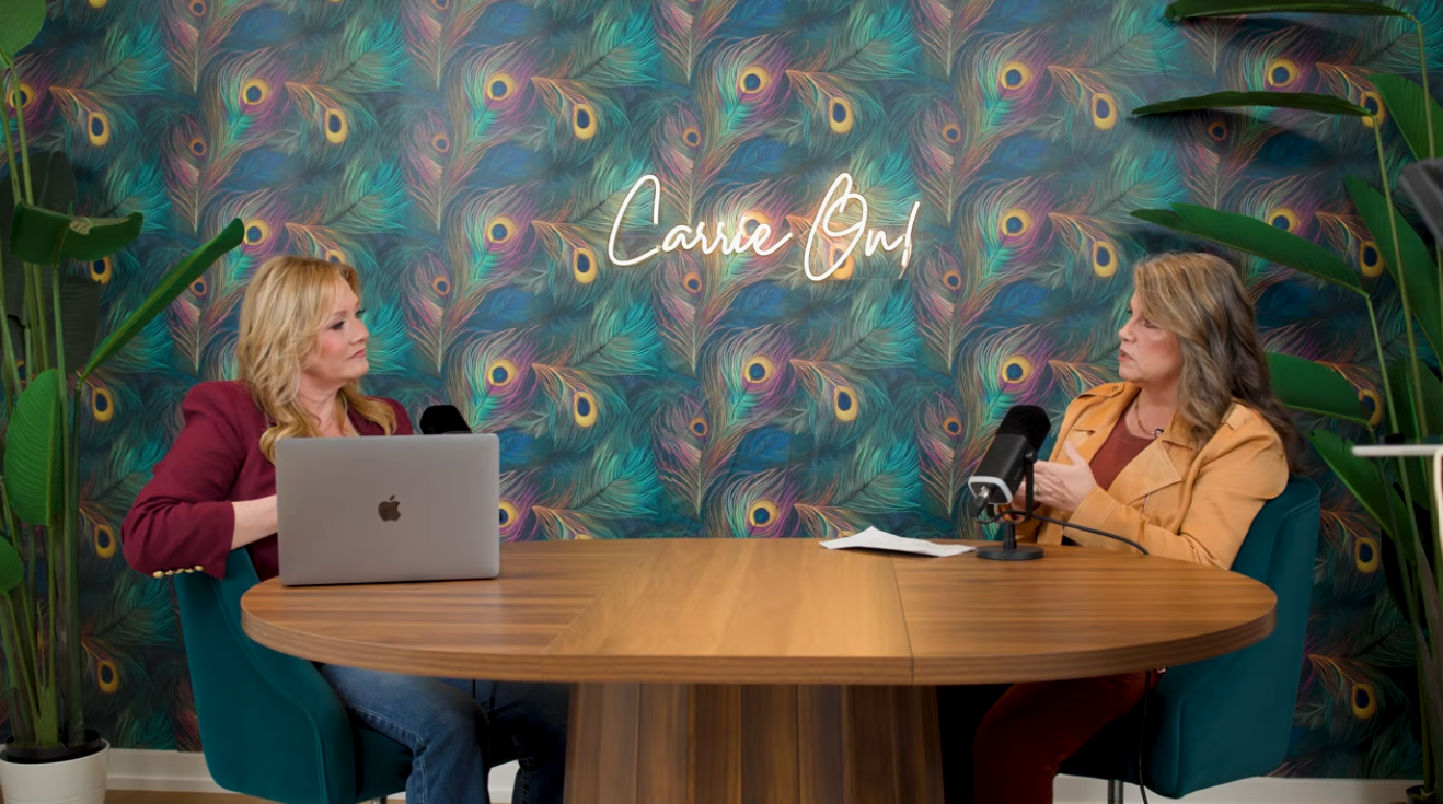 Two women sitting at a wooden table with microphones, one with a MacBook, in front of a colorful peacock feather wallpaper with the text "Caring And"