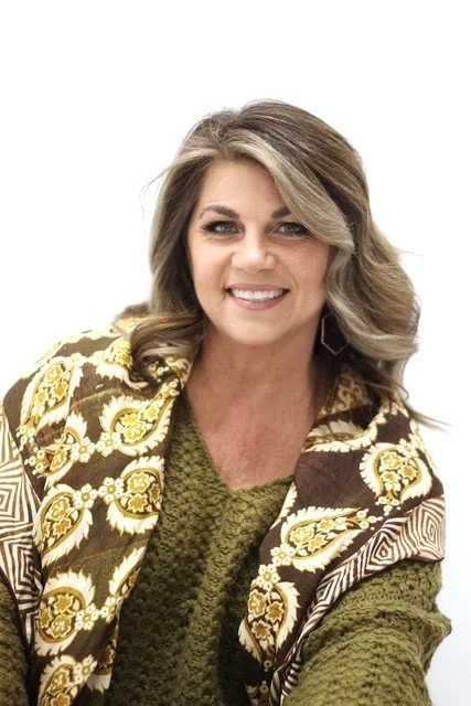 A smiling woman with shoulder-length blonde hair wearing a patterned blazer over a green sweater, against a plain white background.