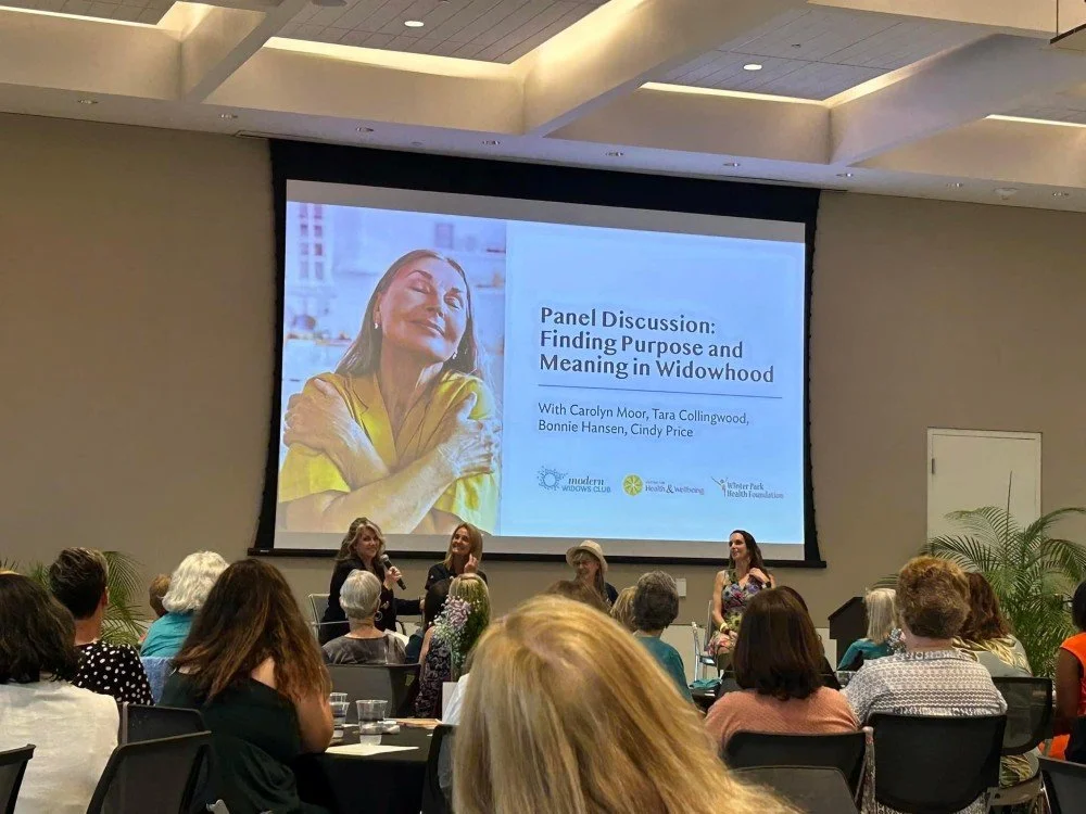 Panel discussion at a conference on finding purpose and meaning in widowhood, featuring four women on stage, with a large screen displaying the discussion topic and participants' names, and audience members seated at tables.