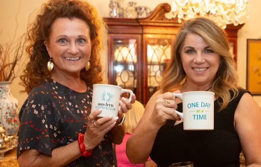 Carolyn and a friend smile, holding coffee mugs that say one day at a time, in a warmly decorated room.