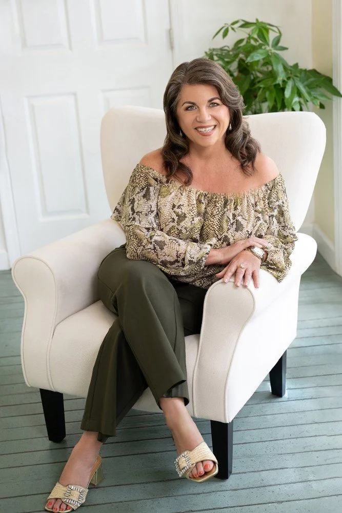A woman with wavy brown hair, wearing an off-the-shoulder snake-print blouse, dark green pants, and embellished open-toe heels, sitting on a white armchair in a well-lit room with a door and plant in the background.