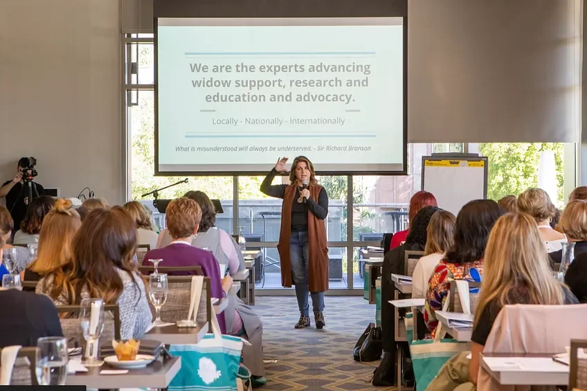 A woman stands in front of a large projection screen giving a presentation to an audience of mostly women in a conference room. The room has large windows with natural light, and some tables have glasses and plates.